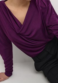 Haut violet à manches longues avec un col en V drapé, fabriqué dans un tissu lisse avec des détails froncés aux épaules. Associé à un pantalon noir.