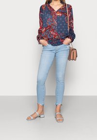 Blusa azul marina con patrón de paisley rojo, mangas largas y puños elásticos, combinada con jeans ajustados de color azul claro y sandalias de tacón bloque azul claro.