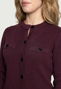 Burgunderfarbener Strickcardigan mit Rundhalsausschnitt, Knopfverschluss und zwei Brusttaschen, ausgestattet mit schwarzen Knöpfen und einem strukturierten Rippenmuster.