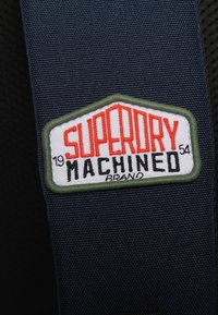 Tejido navy con una superficie texturizada y un parche rectangular. El parche tiene "SUPERDRY" en rojo, "MACHINED" en negro y un borde verde.