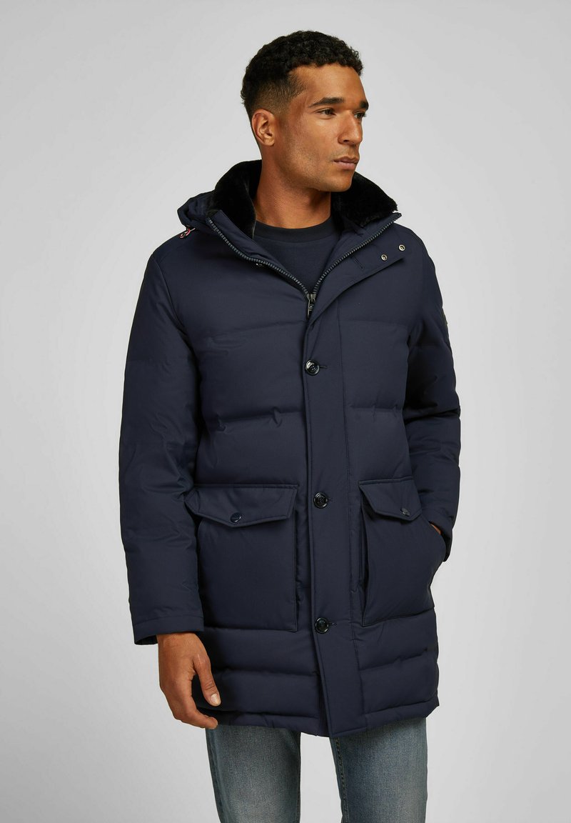 Marineblauwe pufferjas met een rits en knoopsluiting, twee voorzakken en een bontvoering aan de capuchon. Beschikt over een gestikte ontwerp en een gladde textuur.