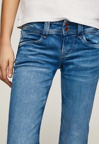 Blå denimjeans med slim fit, sömdetaljer, femfickorsdesign och två bronsknappar vid midjan.