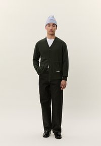 Άνδρας που φοράει σκούρο πράσινο ζακετάκι, λευκό πουκάμισο, μαύρο παντελόνι, μαύρα παπούτσια και ανοιχτό γκρι σκουφάκι, στέκεται μπροστά σε ένα ανοιχτόχρωμο φόντο.