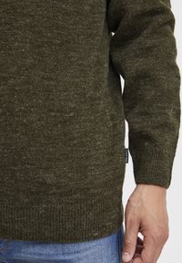 Olivgrüner Strickpullover mit strukturiertem Stoff, gerippten Bündchen und Saum. Verfügt über ein kleines Markenlabel an der Seite.