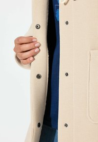 Béžový texturovaný kardigan s tkaným vzorem, vybavený kovovými patentními knoflíky a přední kapsou, vrstvený přes modrou košili.