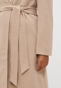 Bata texturizada beige con cinturón de lazo, que presenta un patrón diagonal. Mangas largas y rectas con tela suave visible cerca de la mano.