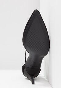 Chaussure noire à talon haut avec un bout pointu et un fin talon aiguille. La tige présente un matériau texturé ; la semelle est lisse avec des rainures pour une meilleure adhérence.