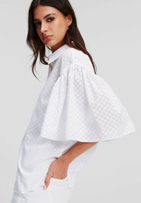 Blusa bianca a maniche corte con un motivo geometrico testurizzato, colletto a punta e vestibilità ampia. Presenta maniche larghe e gonfie.