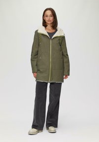 Veste vert olive avec une capuche doublée de fourrure blanche, des fermetures éclair et deux poches avant. Portée avec un pantalon en velours côtelé gris et des chaussures beiges.