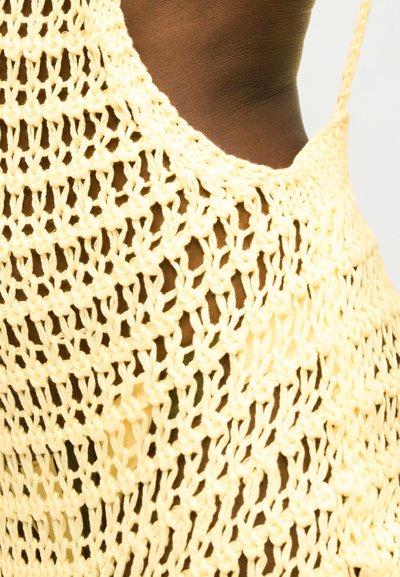 Κοντινό πλάνο σκούρου δέρματος που φοράει ένα παστέλ κίτρινο πλεκτό τοπ χωρίς μανίκια με χαλαρό, ανοιχτό πλέγμα και λεπτό λουράκι στον ώμο.