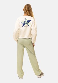 Witte trui met blauw stergrafiek en de tekst "L.A. CITY." Gecombineerd met lichtgroene wijde broekspijpen en grijze sneakers. Textuur van de stof is zichtbaar.