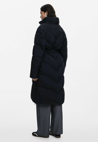 Abrigo de plumón largo y negro con un alto cuello, cintura ceñida y un patrón de acolchado en forma de chevrón. Cuenta con bolsillos laterales y un ajuste holgado.