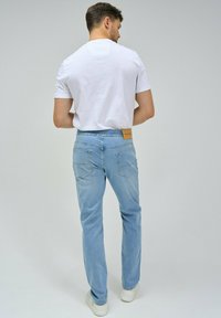 Jeans de mezclilla azul claro con cinturilla elástica, dos bolsillos traseros, costuras en contraste y una etiqueta de cuero marrón en la parte posterior. Camiseta blanca.
