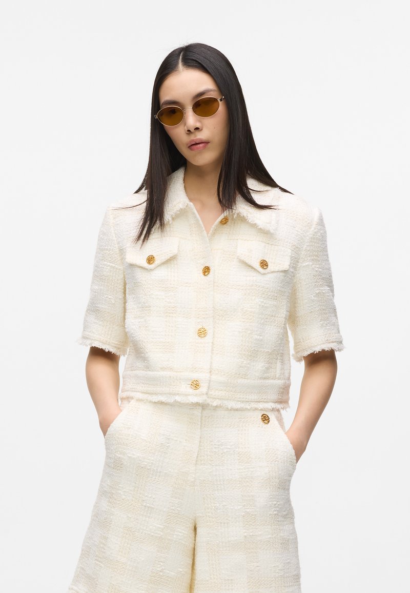 Veste en tweed texturée blanche à manches courtes, ornée de boutons dorés. Associée à un short assorti de couleur claire, créant un look coordonné.