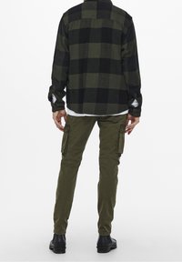 Camisa de franela a cuadros en verde oscuro y negro, combinada con pantalones cargo oliva con bolsillos laterales y botas negras.