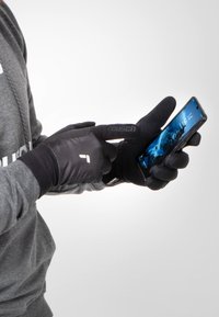 Schwarze Touchscreen-Handschuhe mit einer strukturierten Handfläche, die eng anliegen, ein Logo im Handgelenksbereich haben und verwendet werden, um mit einem Smartphone zu interagieren.