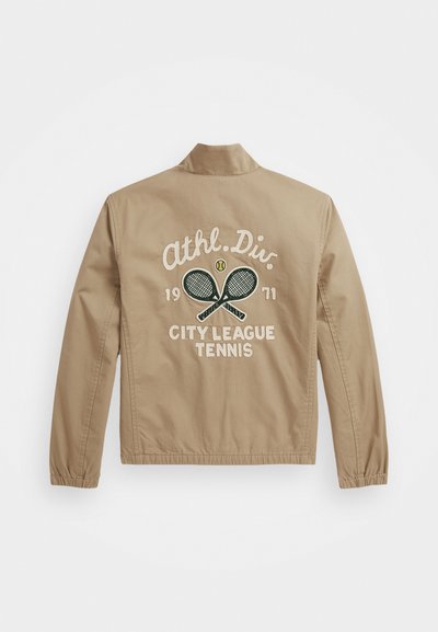 Beige Jacke mit gesticktem Text und Tennisracket-Motiv auf dem Rücken. Elastische Bündchen und ein Stehkragen. Leichter Stoff.