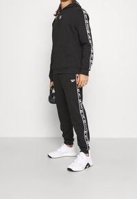 Svart huvtröja och sweatpants med vita sidostreck. Huvtröjan har en framlomma och dragsko; modellen håller en kettlebell. Vita sneakers.