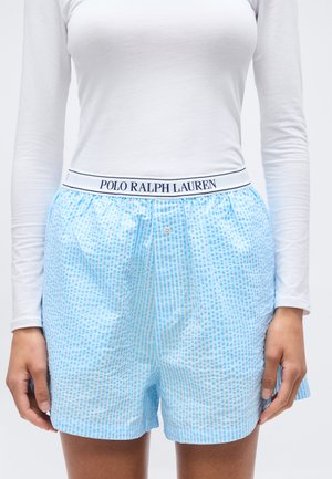 Άτομο που φοράει ανοιχτό μπλε boxer με σχέδιο και λευκή μέση με ετικέτα «POLO RALPH LAUREN» και ένα απλό λευκό μακρυμάνικο πουκάμισο.