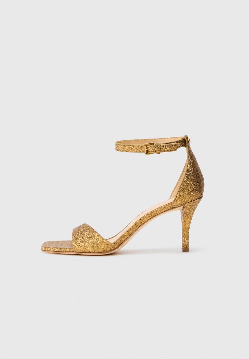 Sandal con tacco alto in glitter oro, con cinturino alla caviglia e fibbia, punta aperta e sottile tacco a stiletto, su uno sfondo neutro.