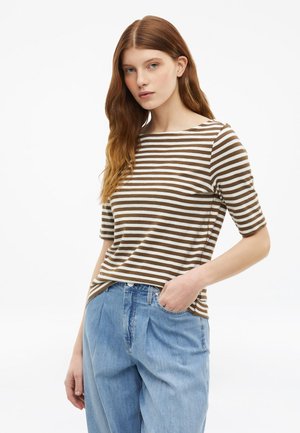 Junge Frau mit langen roten Haaren trägt ein braun-weiß gestreiftes kurzärmliges Oberteil und hellblaue hoch taillierte Jeans.