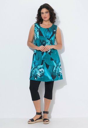 Vrouw met krullend donker haar, gekleed in een mouwloze turquoise tuniek met bloemenprint, zwarte cropped legging en zwarte sandalen met enkelbandje, staand tegen een witte muur.