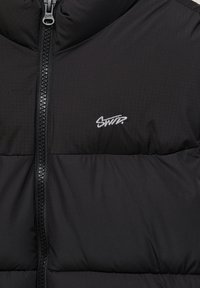 Veste matelassée noire devant avec une fermeture éclair centrale et un petit logo blanc brodé "SWDR" sur la partie gauche de la poitrine.