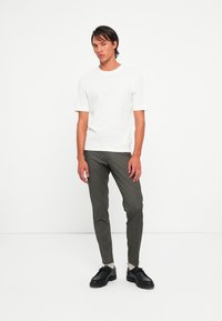 Man standing in a white short-sleeve shirt, gray pants, light socks, and black shoes against a plain white background.