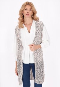 Cardigan senza maniche lavorato a maglia nei toni di rosa pastello, grigio e bianco. Design aperto sul davanti, tessuto testurizzato con motivi sottili, indossato sopra una blouse bianca.
