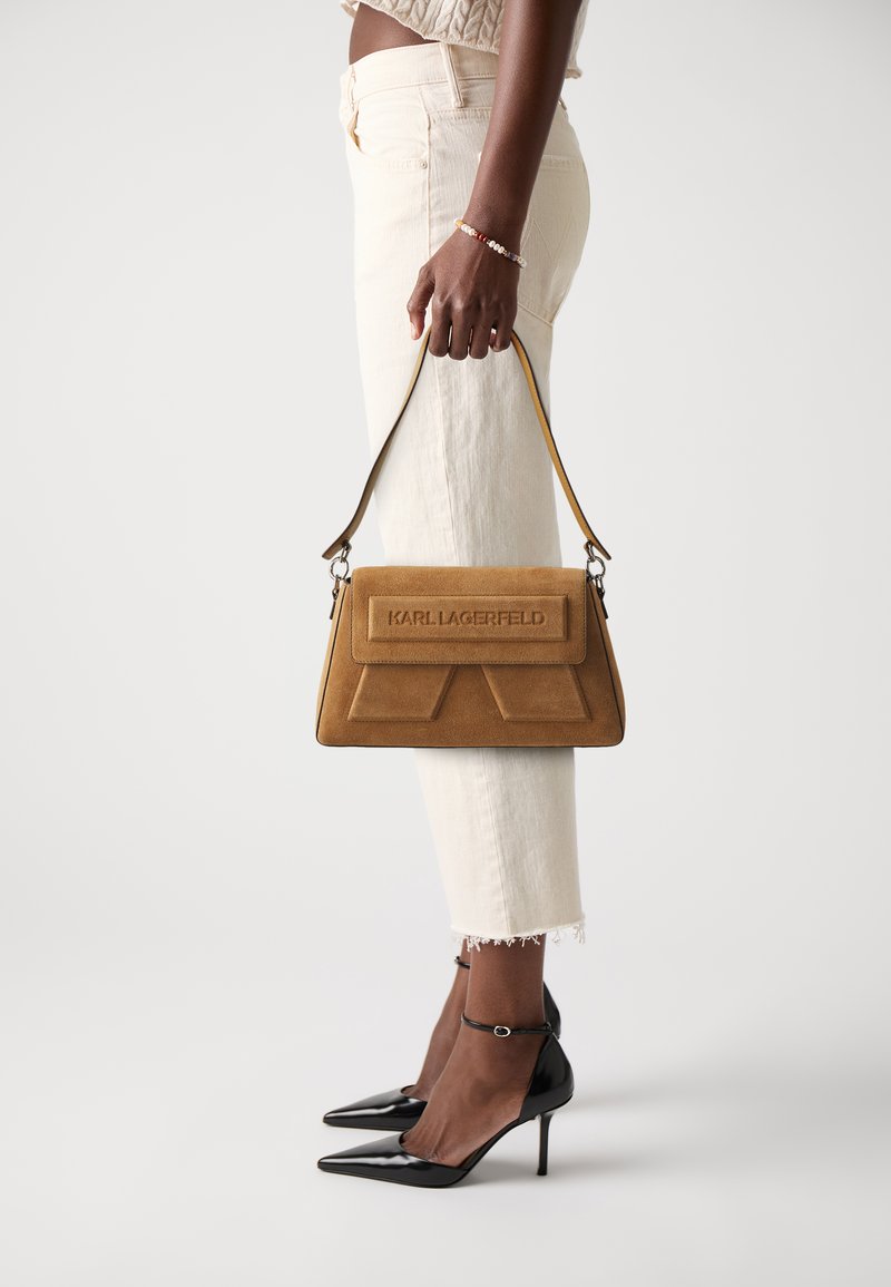 Sac à main en daim marron de forme rectangulaire, avec un logo en relief et une bandoulière amovible, associé à un pantalon blanc court et des escarpins noirs à bouts pointus.