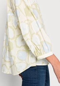 Blouse légère en crème avec un motif floral pastel bleu et vert, présentant une coupe décontractée, des manches bouffantes et des poignets froncés.