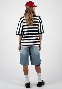 Personne aux longs cheveux portant une casquette rouge, un pull à manches courtes rayé noir et blanc, un short long en jean, des chaussettes blanches et des chaussures noires, debout de dos.