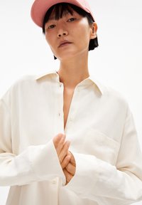 Cremfarbene Hemdbluse mit Kragen, langen Ärmeln und Brusttasche, kombiniert mit einer pinken Kappe. Der Stoff wirkt glatt und leicht.