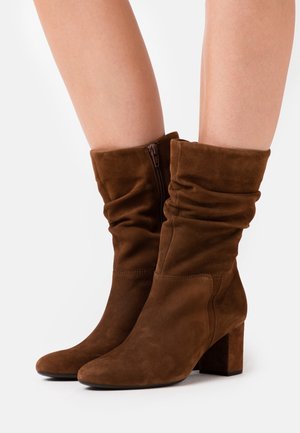 Bottes mi-mollet en suède marron avec design décontracté, fermeture éclair latérale, talons blocs, portées sur jambes nues sur un fond blanc.