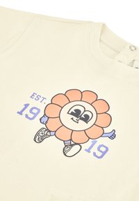 T-shirt in cotone color crema con un personaggio di fiore in stile cartone animato con un viso sorridente, braccia blu e la scritta "EST. 19 19" in viola.