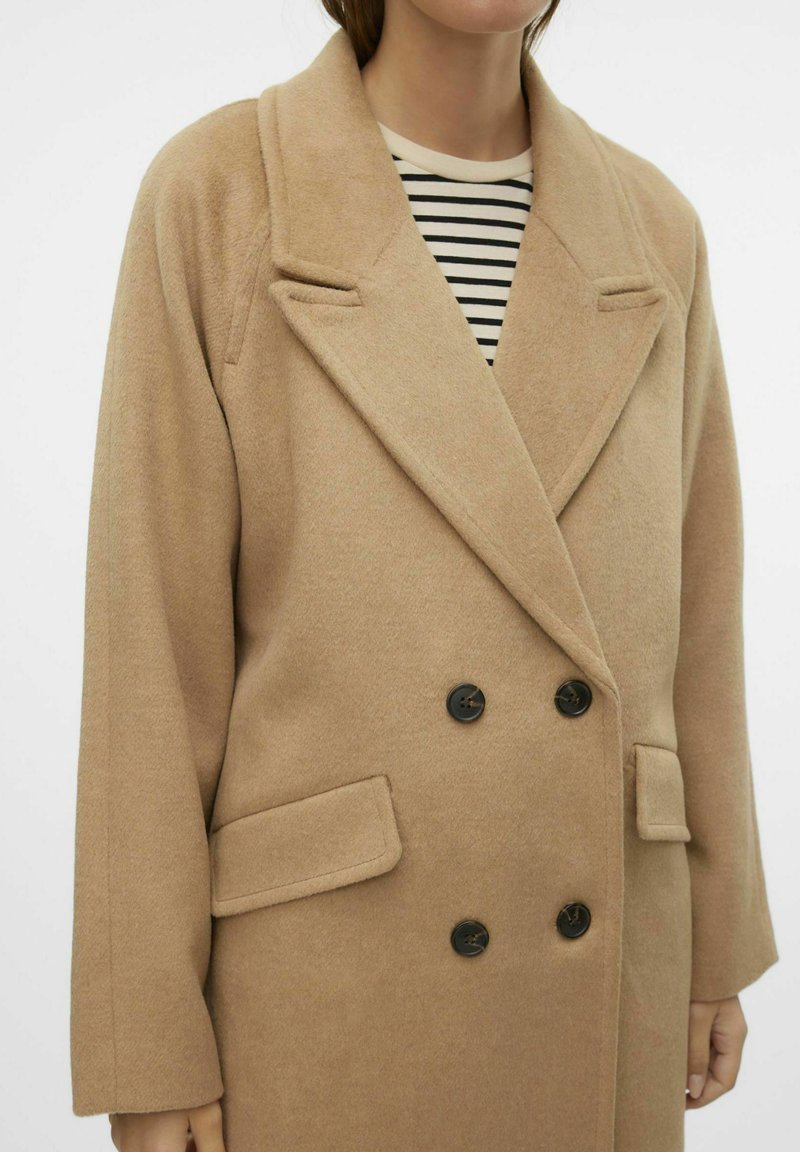 Manteau beige à double boutonnage en tissu doux avec un col à revers, des boutons noirs et des poches latérales, porté sur un haut à rayures.