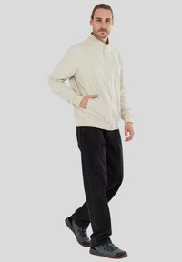 Helle beige Bomberjacke mit hohem Kragen, elastischen Bündchen und zwei Seitentaschen, kombiniert mit schwarzen Jeans und grauen Wanderschuhen.