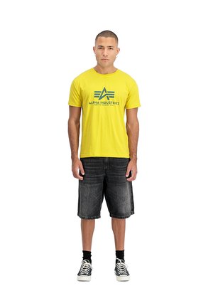 Young man standing facing forward wearing yellow Alpha Industries t-shirt, black denim shorts, black socks, and black sneakers with white laces.