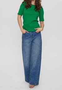 Kvinna med lockigt hår som bär en grön kortärmad topp och vida blå jeans, med händerna i fickorna, stående mot en enkel ljus bakgrund.