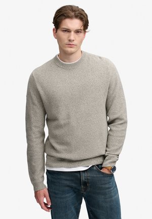 Giovane uomo con capelli castani che indossa un maglione grigio chiaro strutturato, jeans blu e un orologio da polso con cinturino in pelle, in piedi con una mano in tasca.