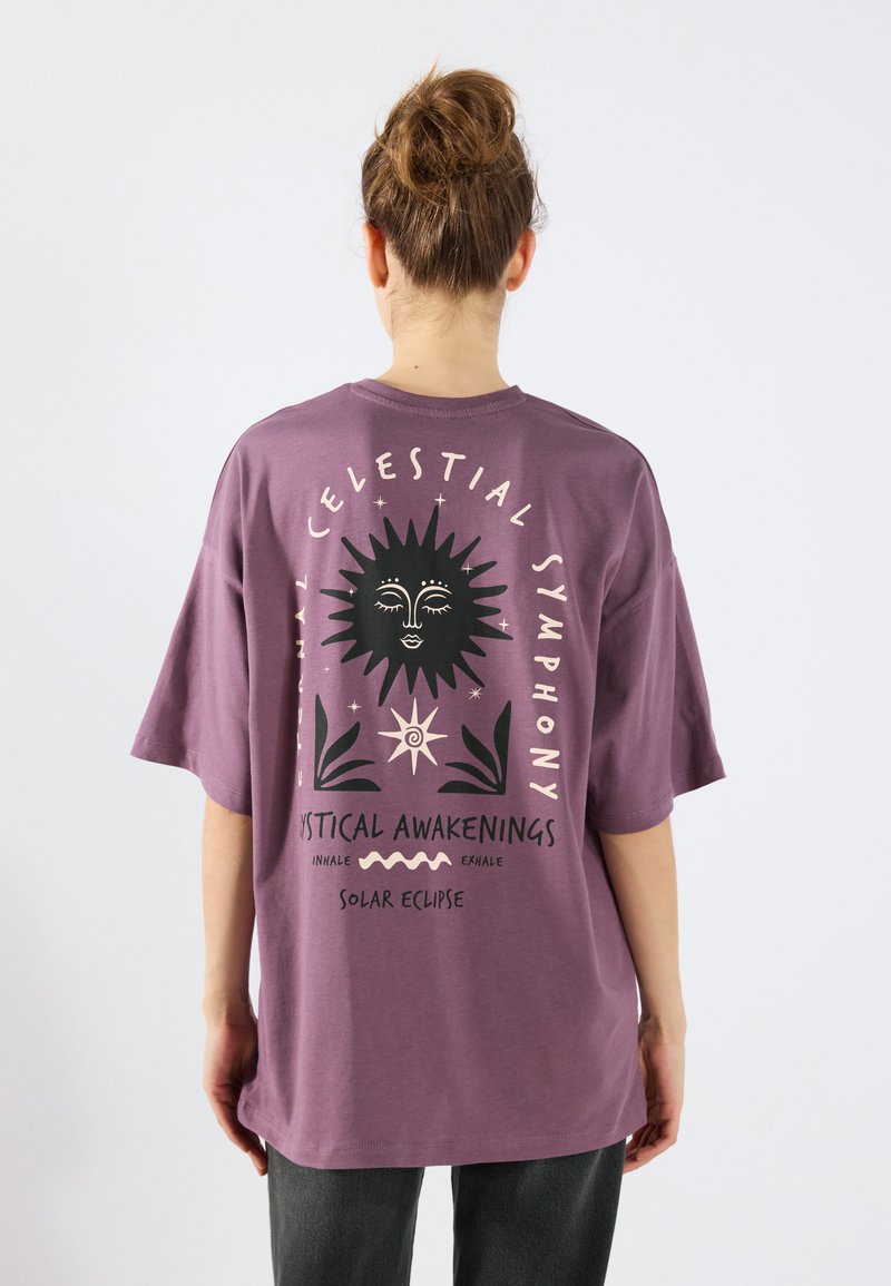 Violetti puuvillapaita lyhyillä hihoilla ja väljän istuvuuden. Selässä grafiikkasuunnittelu, jossa on aurinko, teksti ja kasviaiheet vastakkaisissa väreissä.