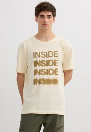 Joven con cabello rizado y oscuro que lleva una camiseta de color crema con la palabra "INSIDE" impresa cuatro veces en marrón, desvaneciéndose hacia la parte inferior.