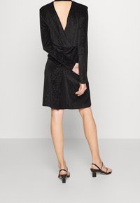 Robe noire à manches longues, dos en V profond, texture douce et accents scintillants. Présente une silhouette ajustée et une coupe genou avec drapés.