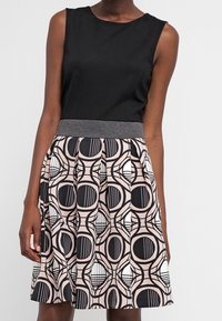 Robe sans manches noire avec une ceinture grise et une jupe plissée arborant un motif circulaire géométrique noir, blanc et rose.