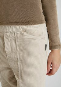 Beige corduroy broek met verticaal geribbelde textuur, voorzien van zijzakken en een contrasterende bruine geribbelde top.
