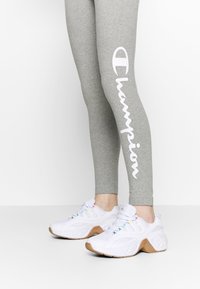 Szare legginsy z białym logo "Champion", zestawione z białymi trampkami z kolorowymi akcentami i gumową podeszwą.