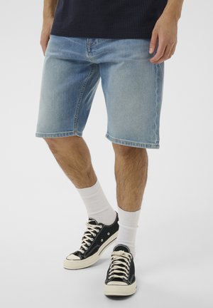 Man draagt lichtblauwe denim shorts, witte sportsokken en zwarte lage sneakers, staand op een effen lichte achtergrond.