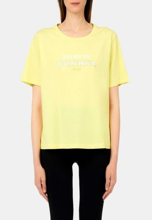 Persona che indossa una maglietta gialla con la scritta "You are my HAPPY HOUR" in bianco, abbinata a pantaloni neri, in piedi contro uno sfondo chiaro.