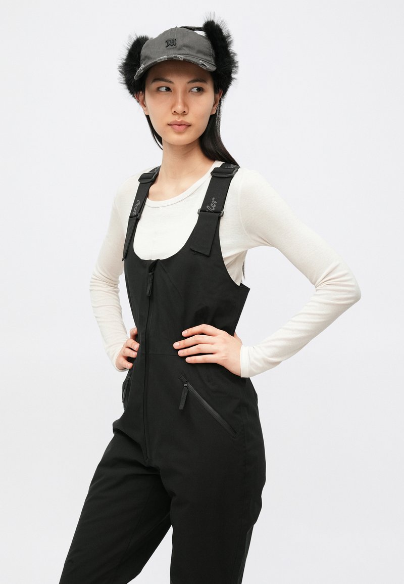 Black overall with zippered pockets and adjustable straps, paired with a gray cap featuring fur accents on the sides.
