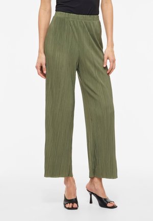 Γυναίκα που φοράει φαρδιά, πλισέ παντελόνια ελιάς με ελαστική μέση, μαύρο αμάνικο τοπ και μαύρα σανδάλια με ψηλό τακούνι και ανοιχτά δάχτυλα.